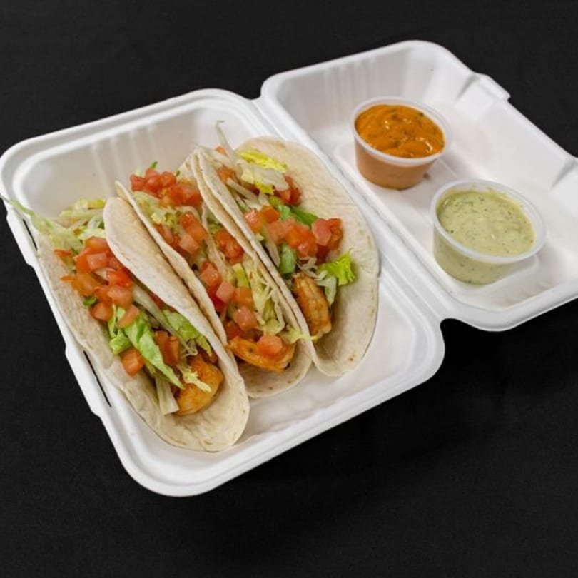 Three shrimp tacos with lettuce and diced tomatoes in a takeout container with two dipping sauces.