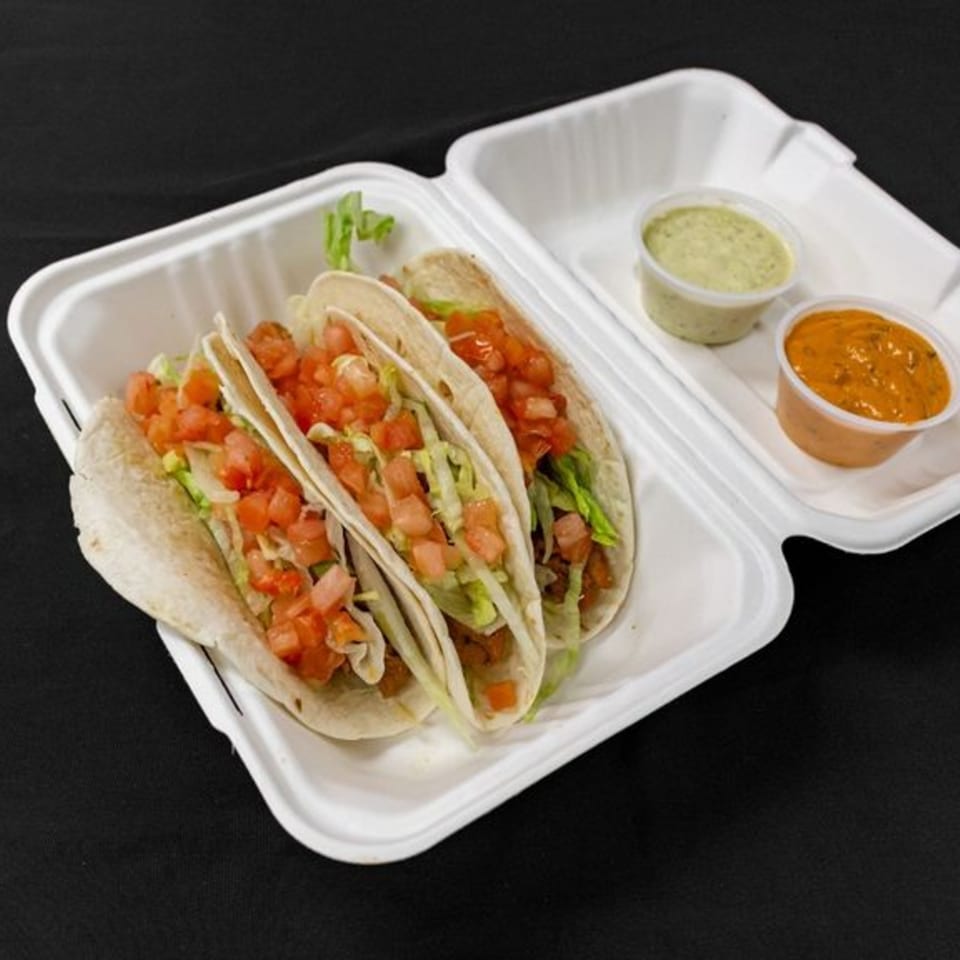 Three fast-food tacos with lettuce and diced tomatoes in a white to-go container with two sauces.