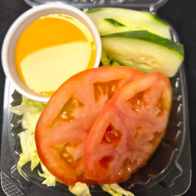 A side salad with sliced tomatoes, cucumbers, lettuce, and a cup of dressing in a plastic container.